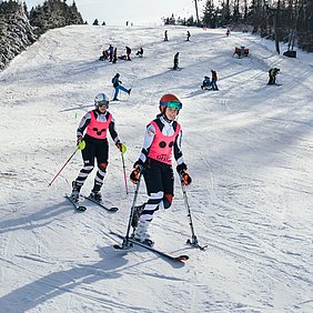 Zwei Skifahrerinnen mit pinken Leibchen fahren die Piste hinab. Die vorderer Skifahrerin fährt auf einem Ski und trägt auf dem Leibchen die Aufschrift Guide.