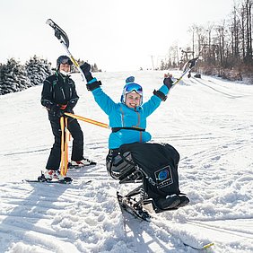 Eine junge Frau sitzt lachend im Monoski und streckt vor Freude beide Arme in die Luft. In den Händen hält sie Ski-Kufen. Hinter dem Monoski steht ihr Begleitläufer und sichert den Ski mit einem Seil. 