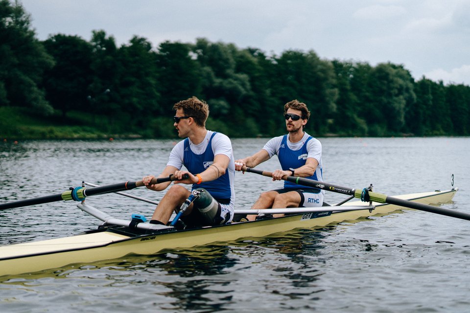 zwei Männer sitzen in einem Ruderboot, ein Mann trägt eine Beinprothese. 