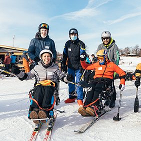 Eine Gruppe lächelt in die Kamera: Links steht eine Biskifahrerin und dahinter ein junger Mann, i der Mitte lacht ein Mann ohne Ski in die Kamera und rechts steht ein Monoskifahrer und dahinter ebenfalls ein junger Mann. 