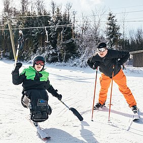 Ein Monoskifahrer und ein stehender Skifahrer stehen im Schnee und lachen in die Kamera. 