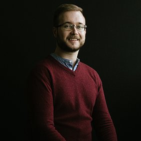 Sebastian sitzt vor einem schwarzen Fotohintergrund und lächelt freundlich in die Kamera. Er trägt eine Brille und einen roten Pullover, darunter ein Hemd.