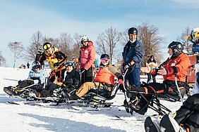 Drei Paare auf Mono- und Biski stehen auf der Piste und hören ihrem Skilehrer zu, der ebenfalls in einem Monoski sitzt. 
